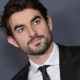Professional headshot: man in a black suit and white shirt, looking to the left against a gray backdrop