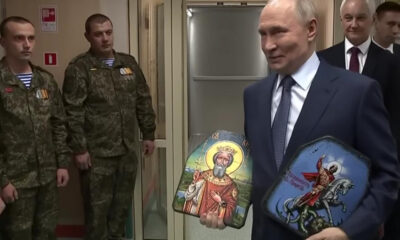 Putin in a suit holds two small religious icons, flanked by soldiers in camouflage and a few officials in the background.