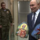 Putin in a suit holds two small religious icons, flanked by soldiers in camouflage and a few officials in the background.