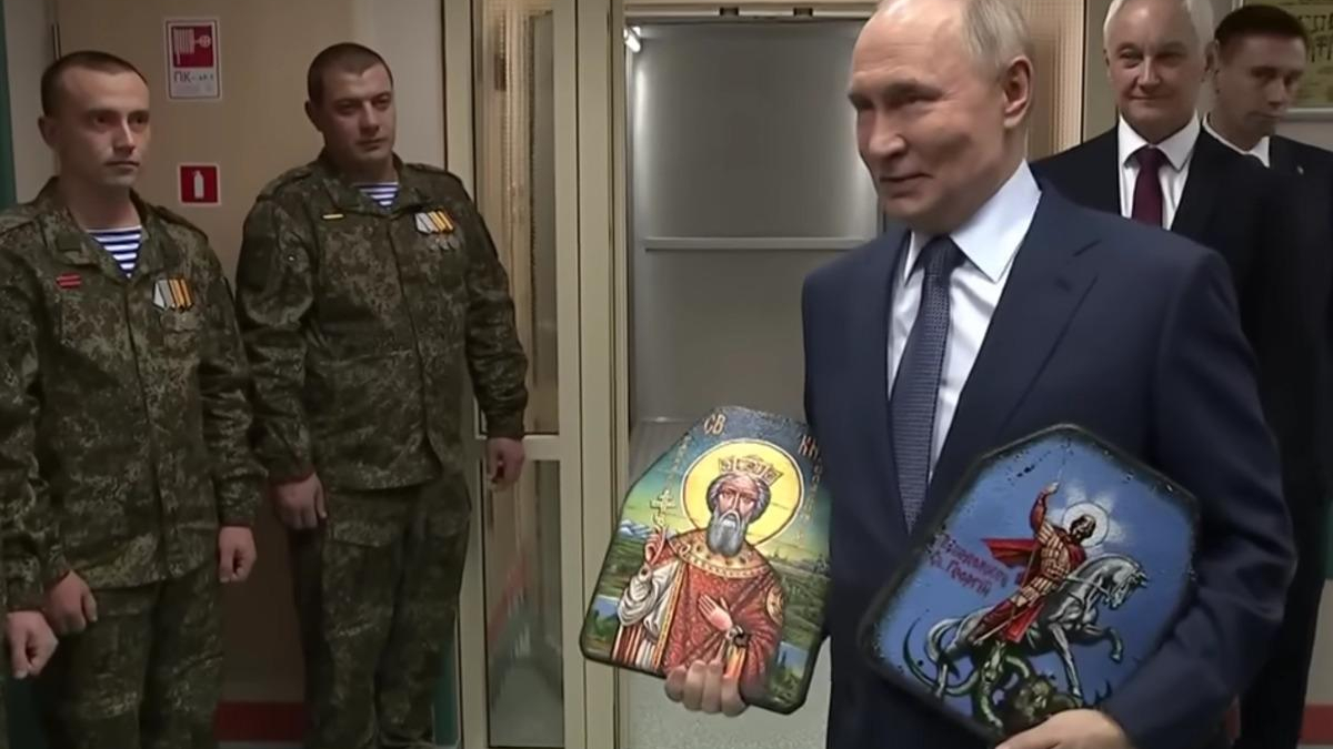Putin in a suit holds two small religious icons, flanked by soldiers in camouflage and a few officials in the background.