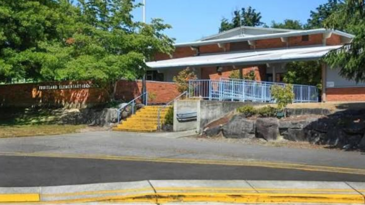 Brick school building with a raised entry, yellow steps, blue railing, and surrounding trees next to a street.
