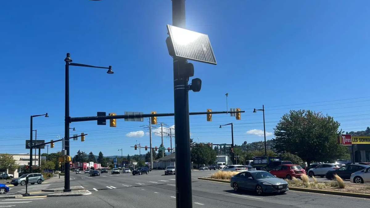 Solar panel mounted on a streetlight at a busy intersection with cars and clear blue sky behind it.