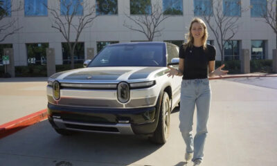 Woman standing beside a white SUV in a parking lot outside a modern office building, gesturing with open hands.
