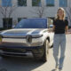 Woman standing beside a white SUV in a parking lot outside a modern office building, gesturing with open hands.