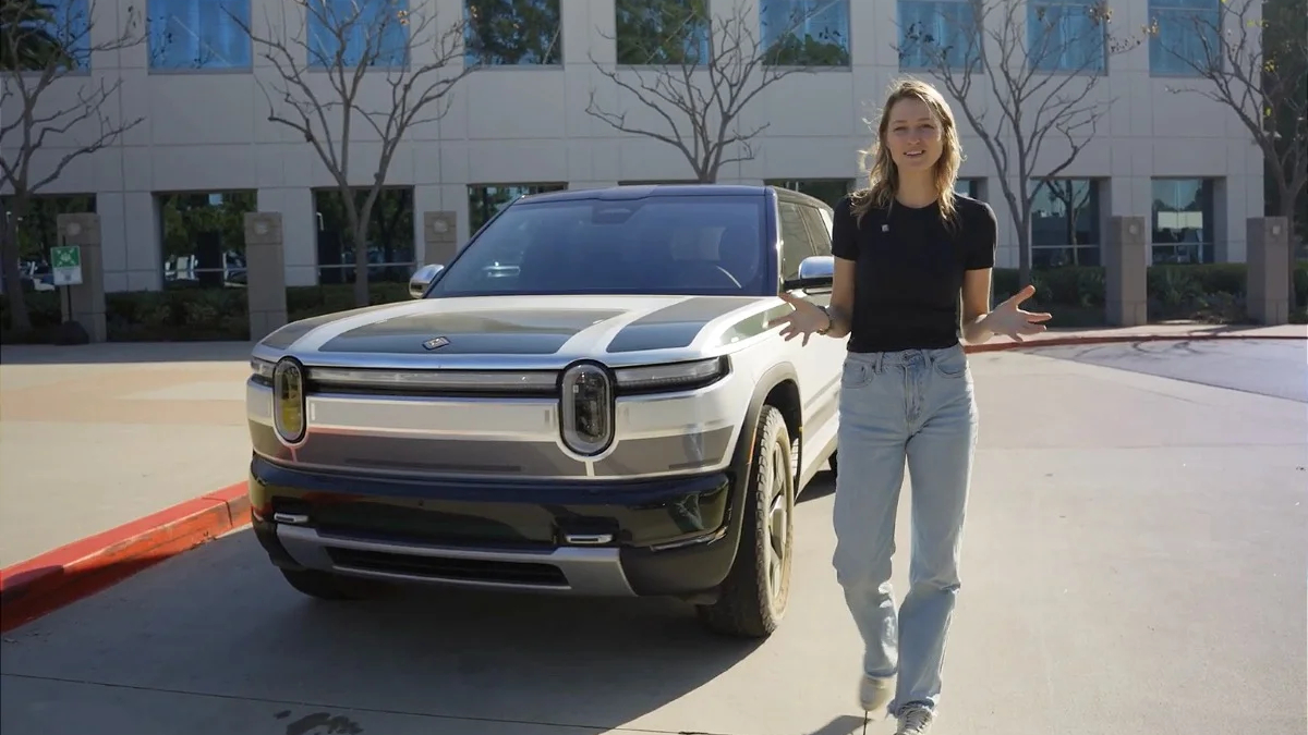 Woman standing beside a white SUV in a parking lot outside a modern office building, gesturing with open hands.