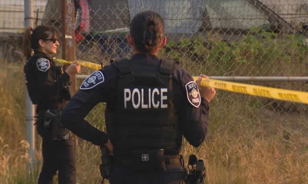 Police officers secure a scene with yellow caution tape, back of officer labeled POLICE visible by a chain-link fence in a grassy area.