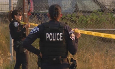 Police officers secure a scene with yellow caution tape, back of officer labeled POLICE visible by a chain-link fence in a grassy area.
