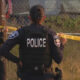 Police officers secure a scene with yellow caution tape, back of officer labeled POLICE visible by a chain-link fence in a grassy area.