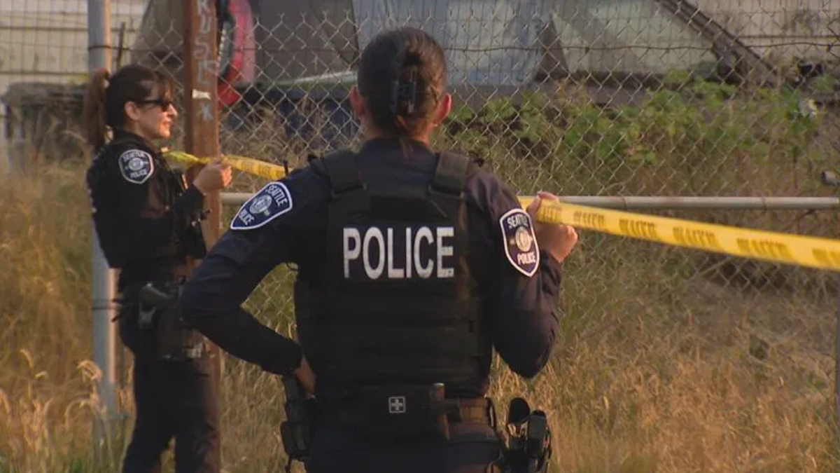 Police officers secure a scene with yellow caution tape, back of officer labeled POLICE visible by a chain-link fence in a grassy area.