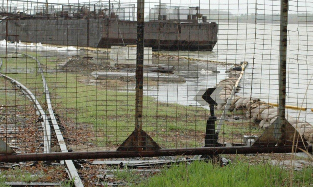 Person in a beige coat leans on a railing at a waterfront jetty, with a rusted pipe and barge in the background behind a chain-link fence.