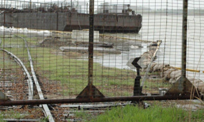 Person in a beige coat leans on a railing at a waterfront jetty, with a rusted pipe and barge in the background behind a chain-link fence.