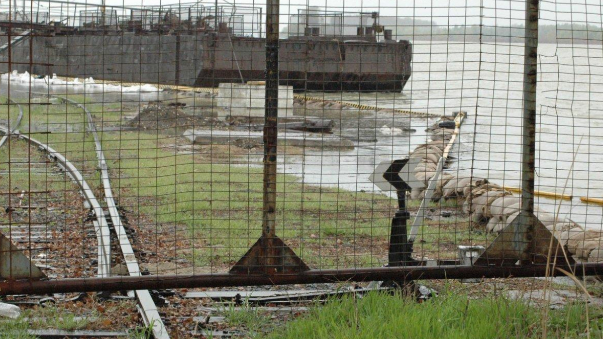 Person in a beige coat leans on a railing at a waterfront jetty, with a rusted pipe and barge in the background behind a chain-link fence.