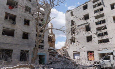 Ruined residential buildings with blown-out windows and rubble, a leafless tree in the foreground, and a damaged vehicle amid debris.