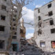 Ruined residential buildings with blown-out windows and rubble, a leafless tree in the foreground, and a damaged vehicle amid debris.