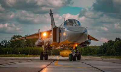 Front view of a silver military fighter jet on a runway with landing gear down and a yellow external tank underneath, against a cloudy sky.