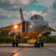 Front view of a silver military fighter jet on a runway with landing gear down and a yellow external tank underneath, against a cloudy sky.