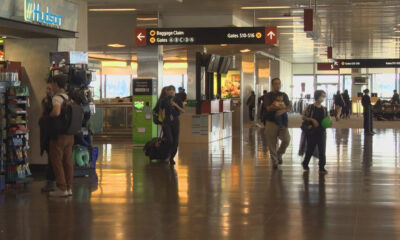 Busy airport terminal with signs for baggage claim and gates, people walking with luggage and shopping area on the left.