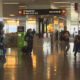 Busy airport terminal with signs for baggage claim and gates, people walking with luggage and shopping area on the left.