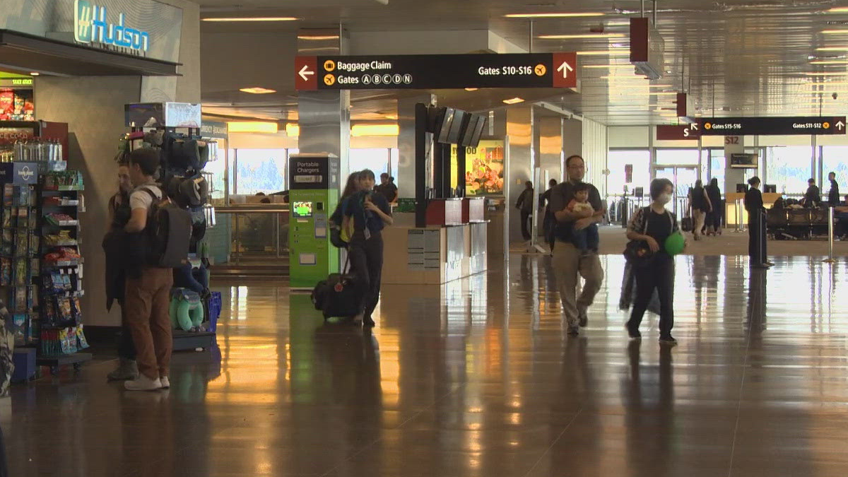Busy airport terminal with signs for baggage claim and gates, people walking with luggage and shopping area on the left.
