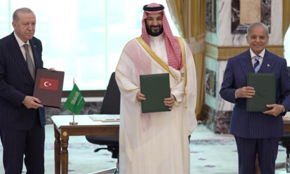 Three men in a signing ceremony hold green folders; Turkish flag visible on the desk and a Saudi official in traditional attire stands center.