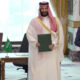 Three men in a signing ceremony hold green folders; Turkish flag visible on the desk and a Saudi official in traditional attire stands center.