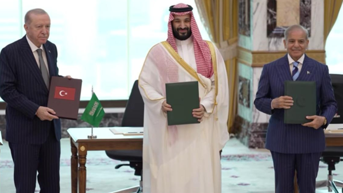 Three men in a signing ceremony hold green folders; Turkish flag visible on the desk and a Saudi official in traditional attire stands center.