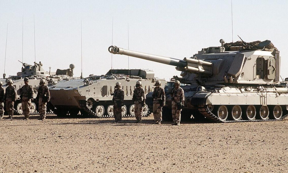 Line of armored tanks with soldiers standing in desert terrain.