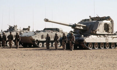 Line of armored tanks with soldiers standing in desert terrain.
