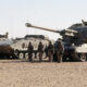 Line of armored tanks with soldiers standing in desert terrain.