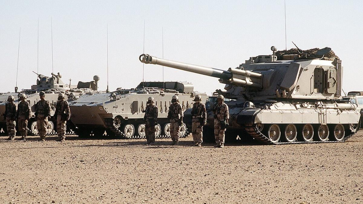 Line of armored tanks with soldiers standing in desert terrain.