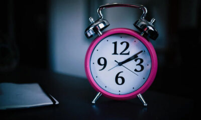 Bright pink analog alarm clock with chrome bells sits on a dark desk beside a small notebook.