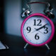 Bright pink analog alarm clock with chrome bells sits on a dark desk beside a small notebook.