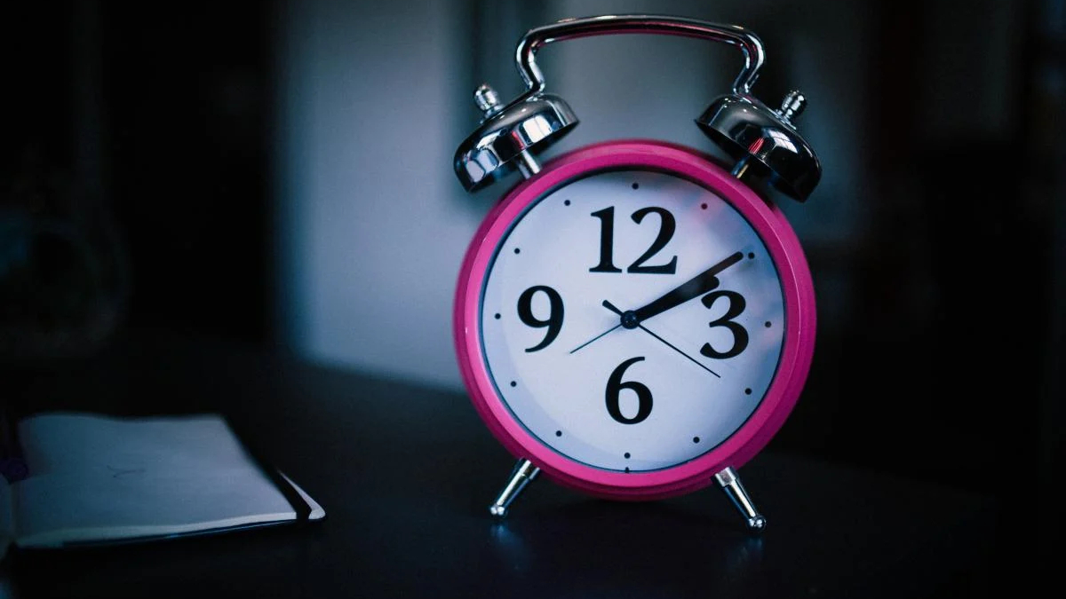Bright pink analog alarm clock with chrome bells sits on a dark desk beside a small notebook.