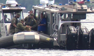 Coast guard officers on a rigid-hull inflatable boat transfer a sealed box to a teammate beside a larger patrol boat on calm water.