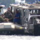 Coast guard officers on a rigid-hull inflatable boat transfer a sealed box to a teammate beside a larger patrol boat on calm water.