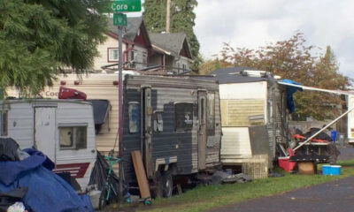Row of old travel trailers and a cluttered camp along a residential street near a Cora St sign.