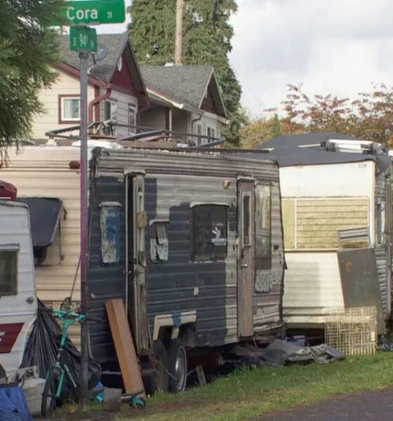 Row of old travel trailers and a cluttered camp along a residential street near a Cora St sign.