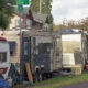 Row of old travel trailers and a cluttered camp along a residential street near a Cora St sign.