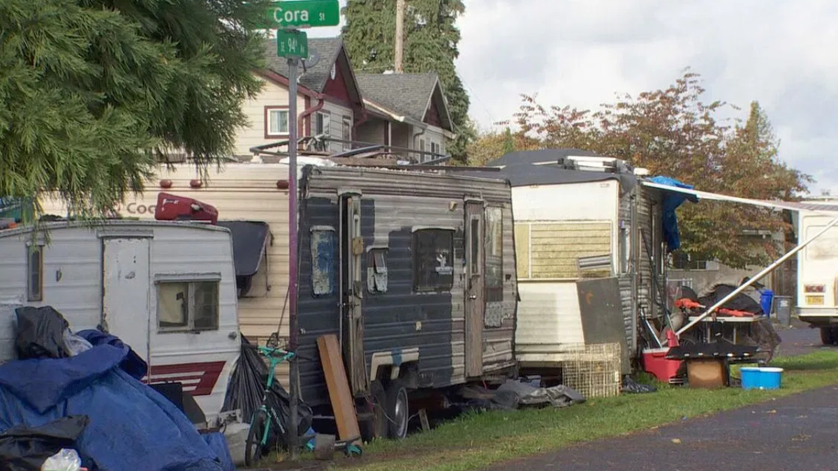 Row of old travel trailers and a cluttered camp along a residential street near a Cora St sign.