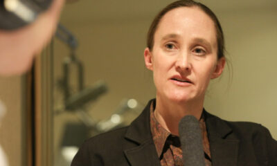 Woman in a dark blazer speaking into a handheld microphone in a studio interview setting
