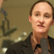 Woman in a dark blazer speaking into a handheld microphone in a studio interview setting