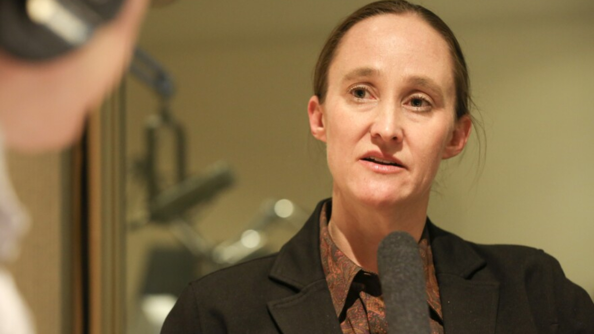 Woman in a dark blazer speaking into a handheld microphone in a studio interview setting
