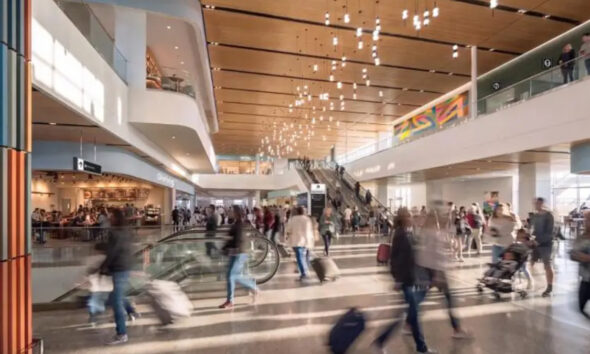 Busy airport terminal with travelers pushing luggage, an escalator, and shops along the concourse.