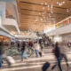 Busy airport terminal with travelers pushing luggage, an escalator, and shops along the concourse.