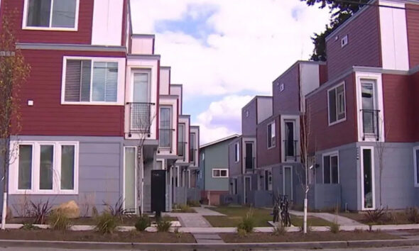 Row of colorful modern townhouses with red and blue siding along a sidewalk, with small trees and shrubs in front.