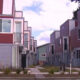 Row of colorful modern townhouses with red and blue siding along a sidewalk, with small trees and shrubs in front.