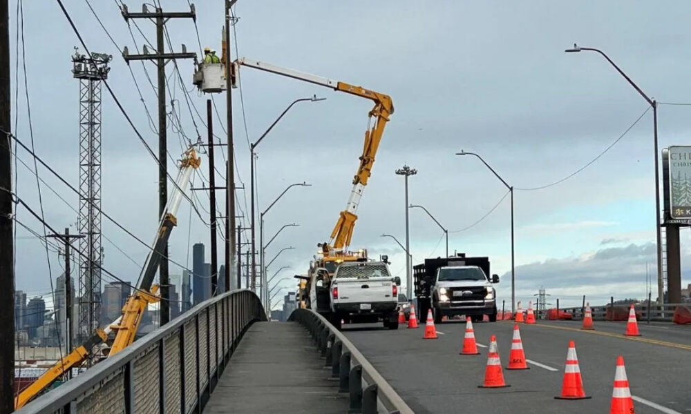 Road construction on a bridge with orange traffic cones, worker in bucket lift repairing power lines, city skyline in the background.