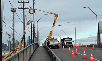 Road construction on a bridge with orange traffic cones, worker in bucket lift repairing power lines, city skyline in the background.