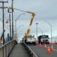 Road construction on a bridge with orange traffic cones, worker in bucket lift repairing power lines, city skyline in the background.