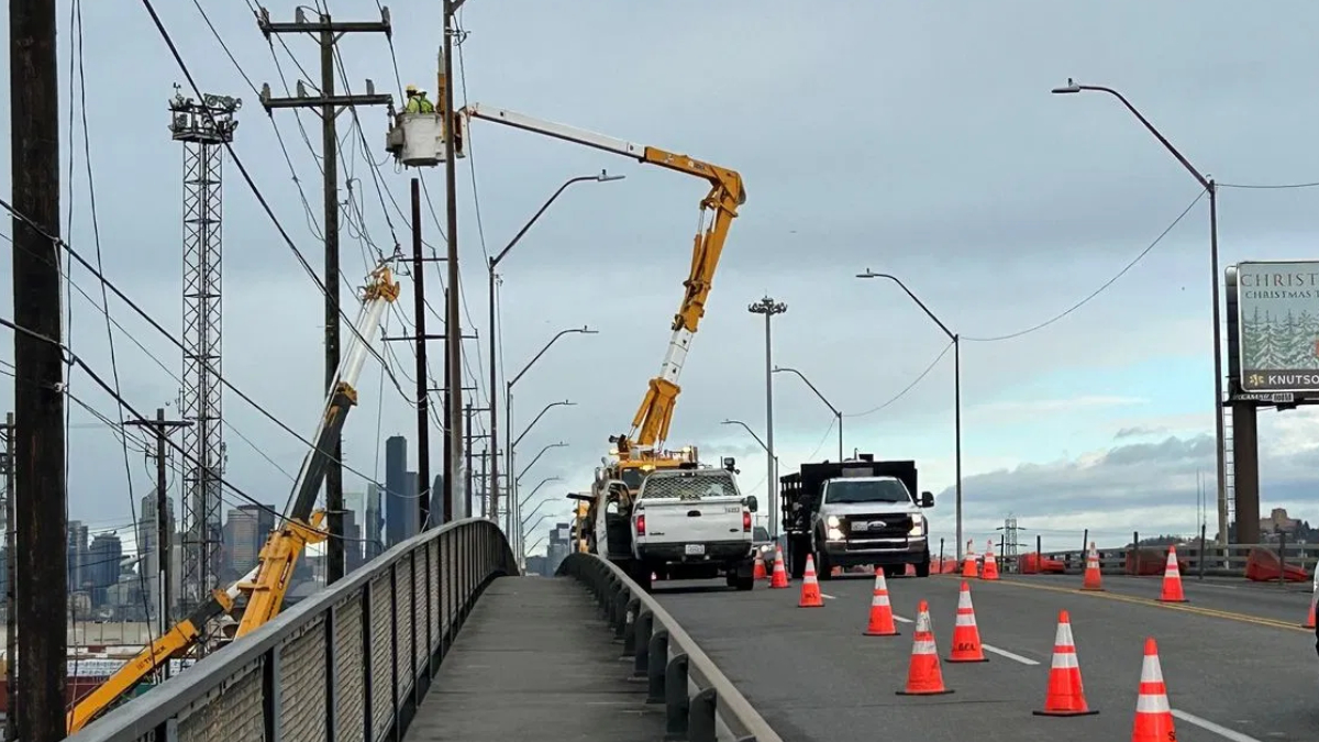 Road construction on a bridge with orange traffic cones, worker in bucket lift repairing power lines, city skyline in the background.